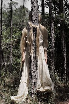 a woman with long hair standing next to a tree in the middle of a forest