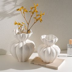 two white vases sitting on top of a table next to a book and yellow flowers