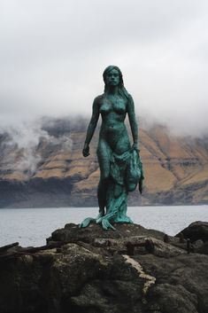 a statue is standing on some rocks by the water with mountains in the back ground