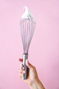 a hand holding a metal whisk over a pink wall with white frosting on it