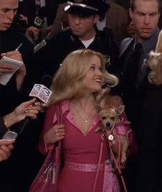 a woman in a pink dress holding a small dog and talking to the media while surrounded by other people