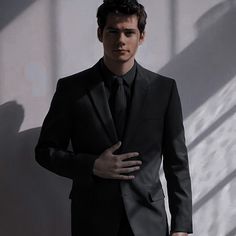 a man in a black suit and tie standing next to a wall with his hands on his chest