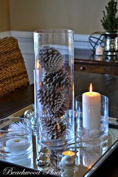 a candle and some pine cones in a glass vase on top of a tray with candles