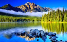 the mountains are covered in clouds and trees near a body of water with rocks on it