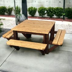 a wooden picnic table with benches in front of it on the sidewalk next to bushes