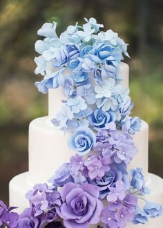 a three tiered white cake with blue flowers on top and purple petals on the bottom