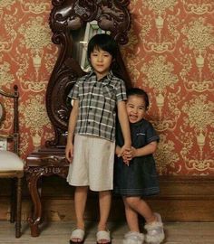 two young children standing next to each other in front of a wallpapered room