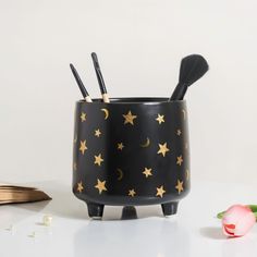 a black ceramic cup with gold stars and moon designs on it, next to a pink rose