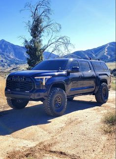 a black truck parked on top of a dirt road next to a tree and mountains