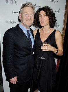 a man and woman standing next to each other in front of a white wall holding wine glasses