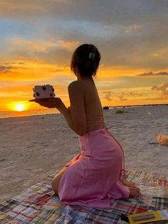 a woman sitting on the beach with a cake in front of her, at sunset