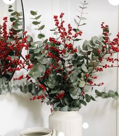 two white vases with red berries and greenery in them on a table next to a mirror