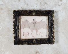 an ornate frame holds a drawing of a skeleton and two white horses on a marble surface