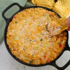 a hand holding a tortilla chip over a skillet filled with chicken and cheese