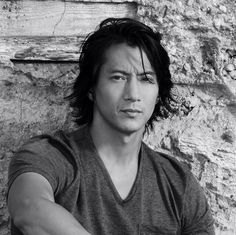 black and white photograph of a man with long hair leaning against a stone wall looking at the camera