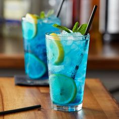 two glasses filled with blue liquid and limes on top of a wooden table next to a black straw