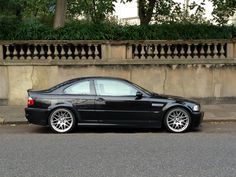 a black car parked on the side of a road next to a wall and trees