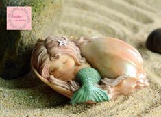 a little mermaid statue laying on top of a sandy beach next to rocks and pebbles