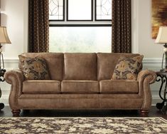 a brown couch sitting in front of a window next to a lamp and rug on top of a wooden floor