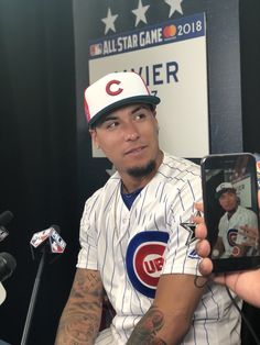 a baseball player holding up a cell phone in front of microphones and an american flag