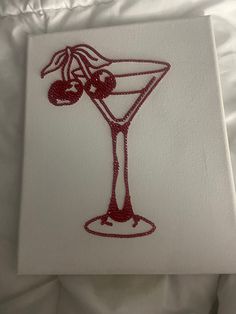 an embroidered cocktail glass with cherries on the rim and in it is white paper