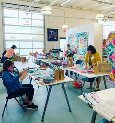 a group of people sitting at tables working on art projects in a room with large windows