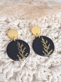 two black and gold earrings sitting on top of a white knitted blanket, one with a plant in it's center