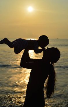 a woman holding a child on her shoulders at the beach as the sun goes down