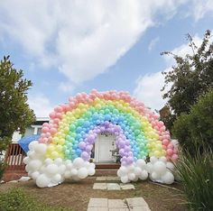 a rainbow arch made out of balloons in front of a house with blue sky and clouds