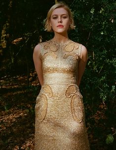 a woman wearing a gold dress standing in front of some bushes and trees with her hands on her hips