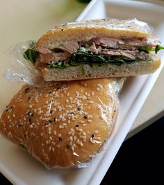 a sandwich with meat, lettuce and sesame seeds wrapped in plastic on a white plate