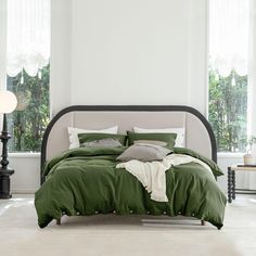 a bed with green sheets and pillows in a white room next to two large windows