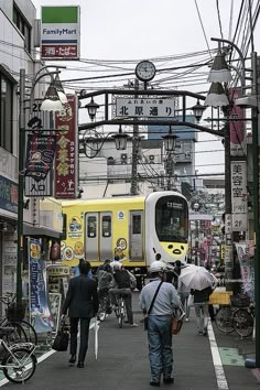 people are walking down the street in front of a yellow and white train that is passing by