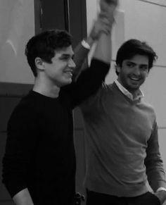 two young men standing next to each other in front of a wall holding up their hands