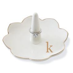 a ring sits on top of a white flower shaped plate with the letter k in gold lettering