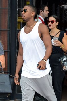 a man in white tank top and grey pants walking down the street with a black handbag