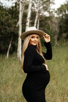 a pregnant woman in a black dress and cowboy hat poses for a photo while standing in tall grass