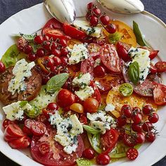 a white plate topped with tomatoes, cheese and other vegetables next to two spoons