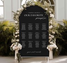 a black and white wedding seating sign with flowers on it, surrounded by greenery