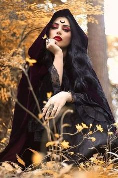 a woman with long black hair and makeup is sitting in the woods