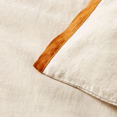 an orange and white pillow on top of a beige linen bed sheet with brown trim