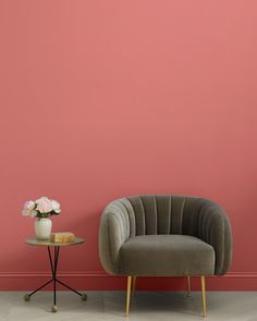 a chair next to a table with a vase on it and a pink wall in the background