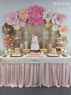 a table topped with lots of cakes and desserts next to a wall covered in paper flowers