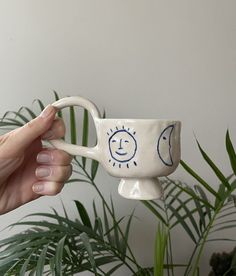 a hand holding a coffee cup with the sun and moon painted on it in front of a potted plant