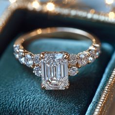 an emerald colored diamond ring sitting on top of a blue velvet box with gold trimmings