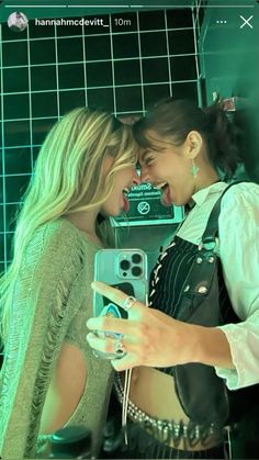 two women standing next to each other in front of a green tiled wall and one holding a cell phone