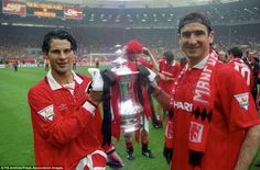 two men in red shirts holding up a silver trophy on a field with other people