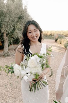 a woman in a white dress holding a bouquet of flowers and smiling at the camera