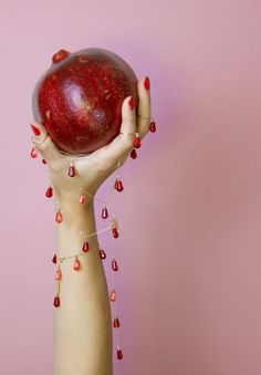 a woman holding an apple up to her face in front of a pink background with beads