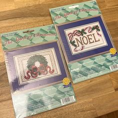 two cross - stitch christmas cards on a wooden table, one with a wreath and the other with an ornament